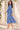 A woman wearing a blue and white paisley midi dress with long sleeves and a tie waist, walking outdoors in front of a beige stucco wall with shadows and blue flowers.