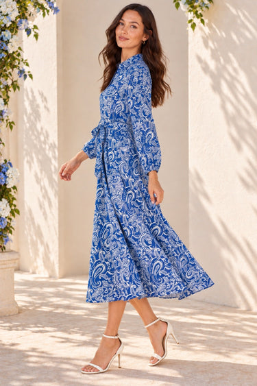 A woman wearing a blue and white paisley midi dress with long sleeves and a tie waist, walking outdoors in front of a beige stucco wall with shadows and blue flowers.