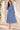 A woman models a blue and white paisley midi dress with long sleeves and a matching waist tie, standing in a sunlit outdoor setting with a cream-colored wall and shadows.