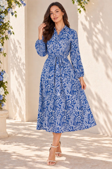 A woman models a blue and white paisley midi dress with long sleeves and a matching waist tie, standing in a sunlit outdoor setting with a cream-colored wall and shadows.